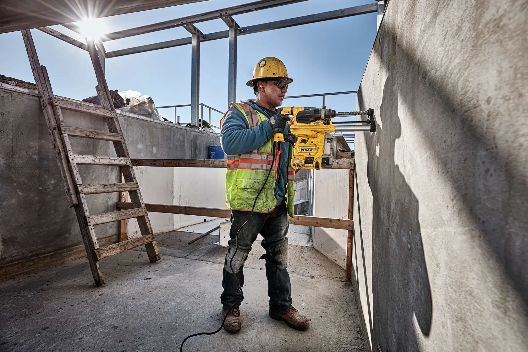 Un trabajador de la construcción con casco de seguridad y chaleco de alta visibilidad opera un taladro rotativo amarillo DEWALT, modelo D25333K, para perforar un agujero en una pared de concreto en un sitio de construcción al aire libre. El trabajador está de pie cerca de una escalera de madera bajo una estructura parcialmente construida, con la luz del sol entrando.