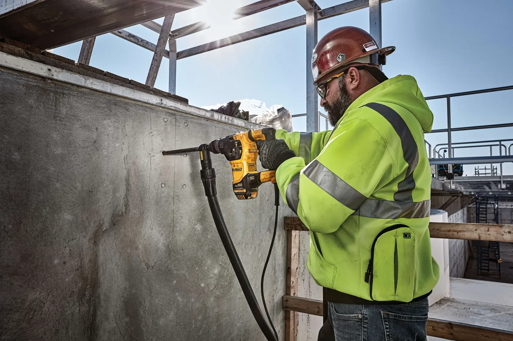 Una persona con casco de seguridad y chaqueta de alta visibilidad está usando una herramienta eléctrica amarilla DEWALT para perforar una pared de concreto en un sitio de construcción.