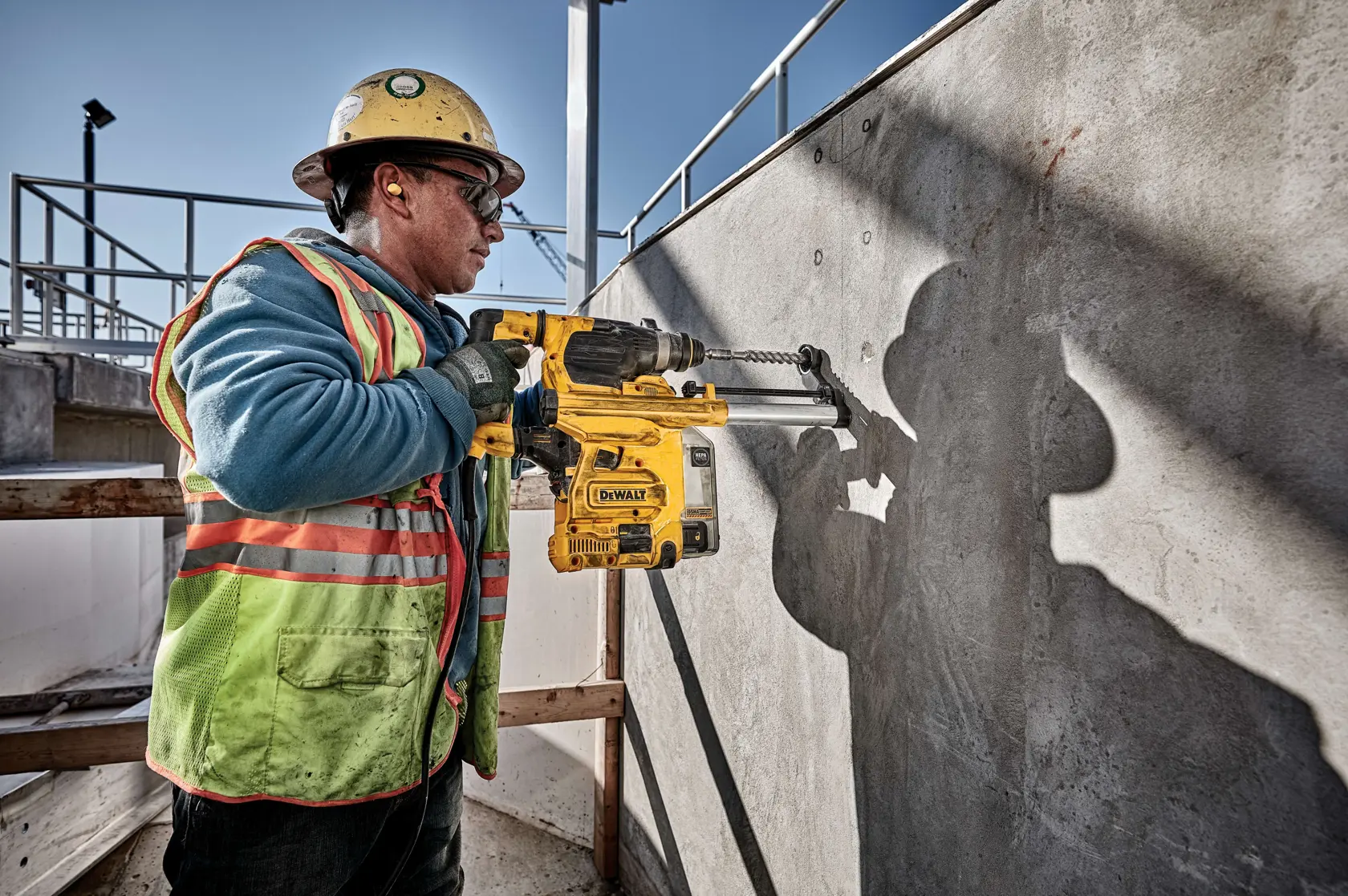 Un trabajador de la construcción con equipo de seguridad opera un taladro rotatorio DEWALT D25333K contra una pared de hormigón en una obra al aire libre.