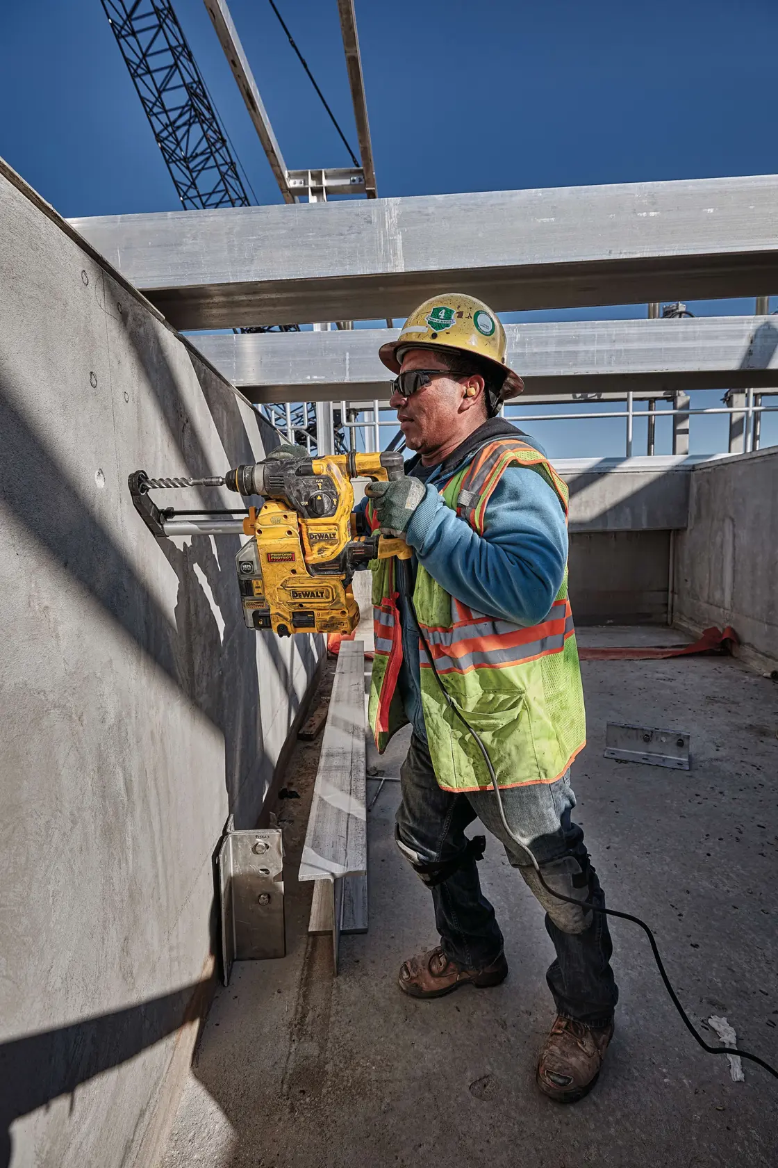 Un trabajador de la construcción con chaleco de seguridad y casco está usando un martillo perforador rotativo DEWALT D25333K para perforar una pared de concreto en una obra. La cara del trabajador está desenfocada para proteger su privacidad.