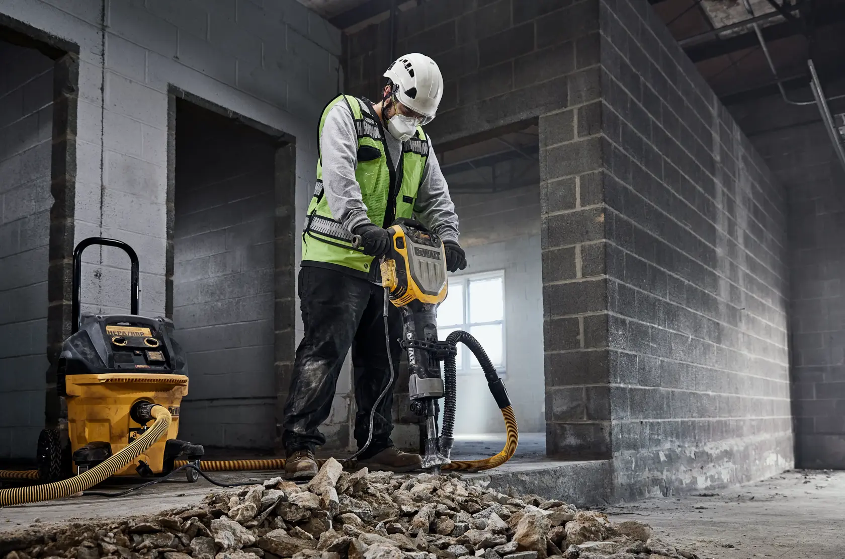 Un travailleur de la construction portant un gilet de sécurité et un casque utilise un marteau-piqueur DEWALT (modèle D25966) pour casser du béton à l'intérieur d'un bâtiment en construction. Un extracteur de poussière DEWALT est connecté au marteau-piqueur, et des gravats de béton cassé sont éparpillés sur le sol.