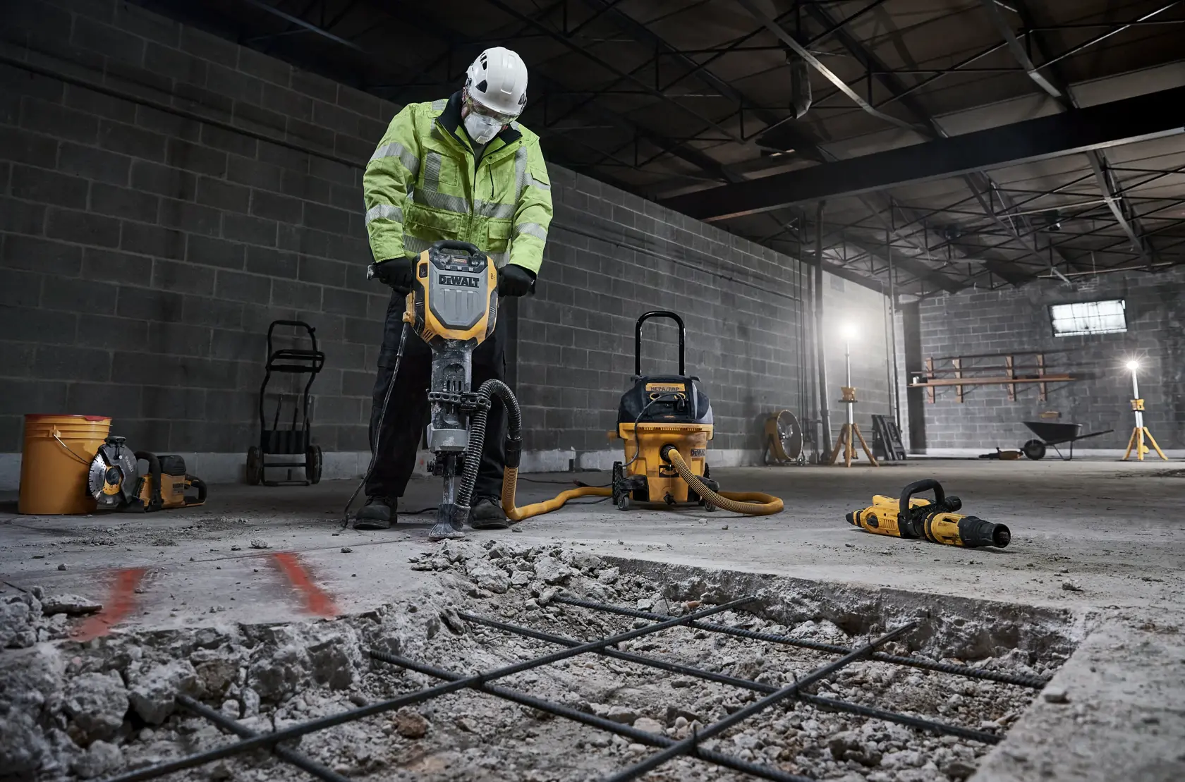 Un travailleur portant un casque de sécurité et une veste haute visibilité utilise un marteau démolisseur DEWALT pour casser du béton dans un environnement industriel. Divers outils et équipements de construction, dont un aspirateur et des outils électriques, sont visibles en arrière-plan.