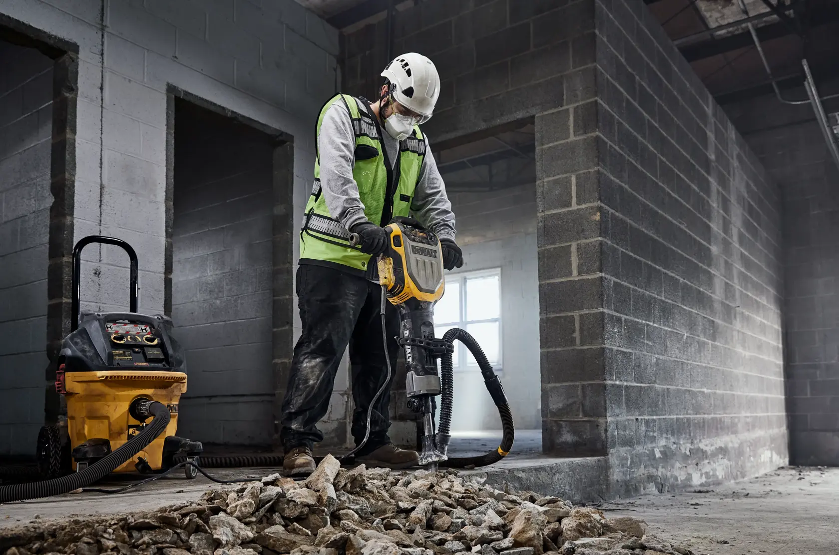 Un ouvrier du bâtiment portant un casque blanc et un gilet de sécurité vert utilise un marteau-piqueur DEWALT pour casser du béton et des pierres à l'intérieur d'un bâtiment en construction. À proximité, un aspirateur jaune DEWALT est connecté au marteau-piqueur pour l'extraction de la poussière.