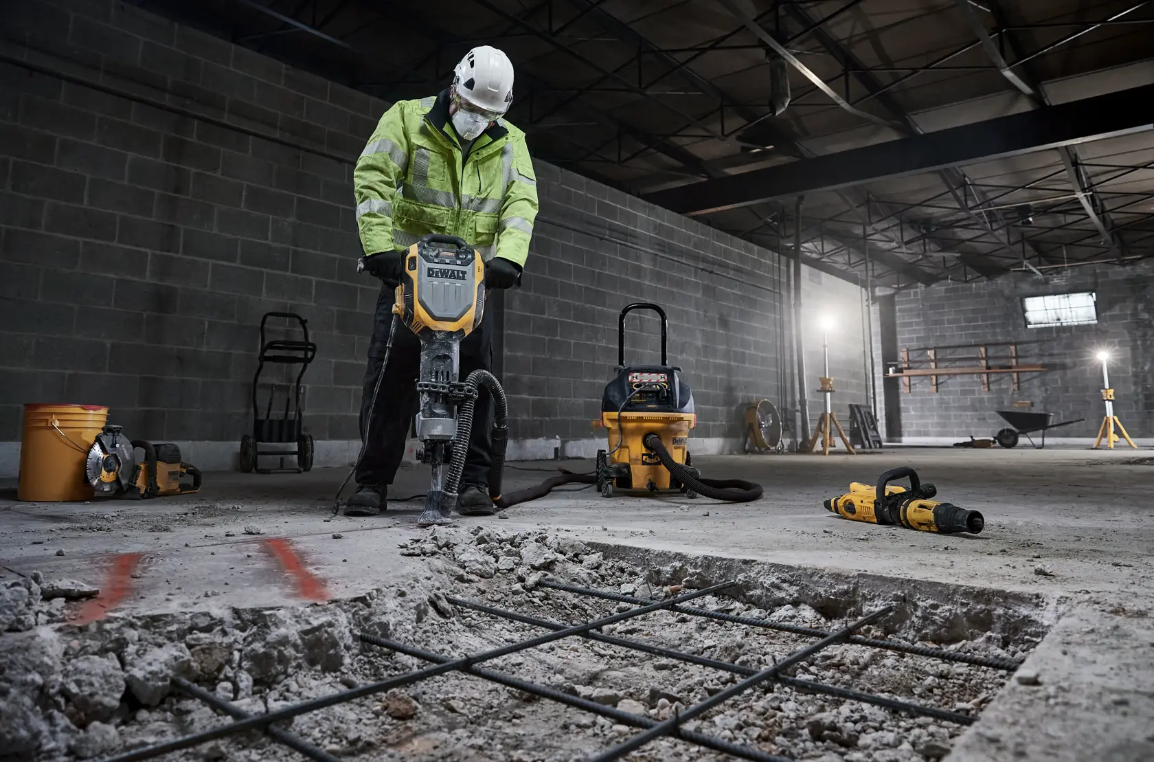 Un ouvrier du bâtiment utilise un marteau piqueur DEWALT D25966 pour casser du béton dans un environnement industriel intérieur. L’ouvrier porte un casque de sécurité et une veste haute visibilité. D’autres outils et équipements DEWALT, tels qu’un aspirateur, des perceuses et un seau, sont visibles en arrière-plan.