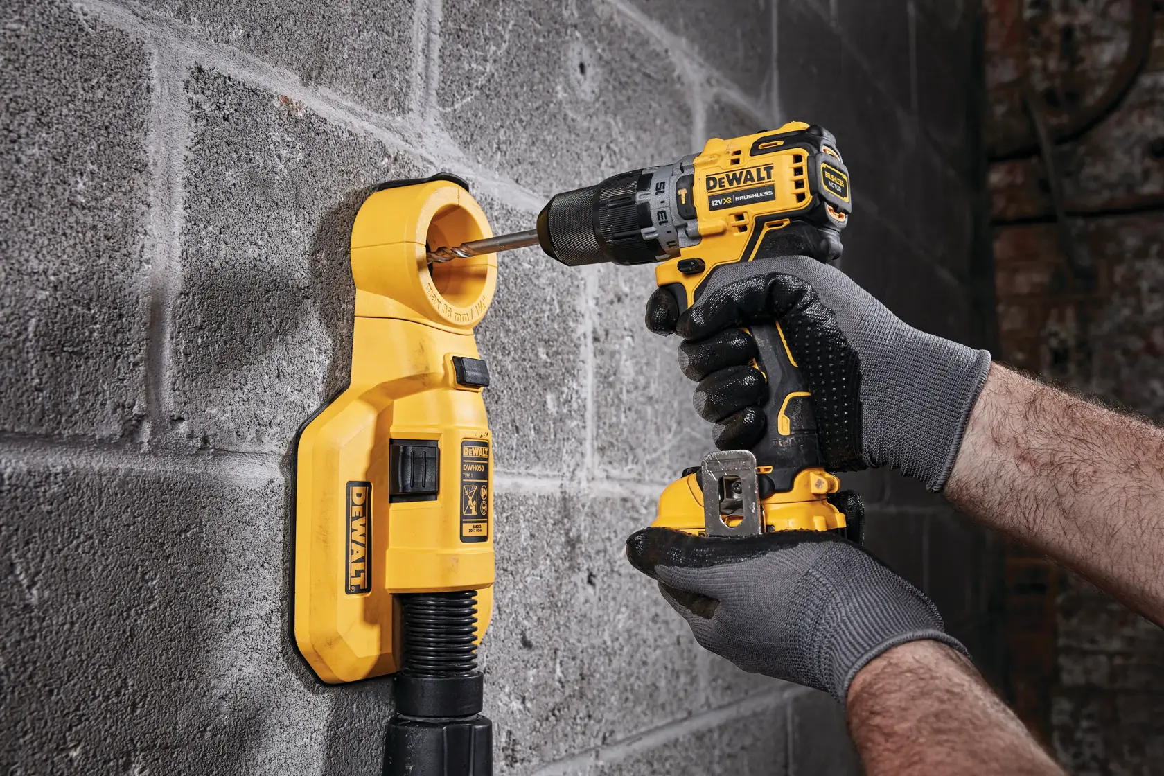 Eine Person mit grauen Handschuhen verwendet einen gelben kabellosen DEWALT-Bohrer, um in eine Betonwand zu bohren, wobei ein gelber Staubabsauger zum Sammeln von Schmutz angebracht ist.
