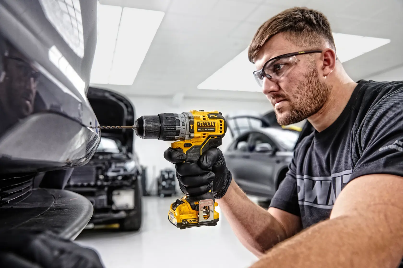 Eine Person mit schwarzen Handschuhen verwendet einen gelben DEWALT DCD706D2 Akku-Bohrschrauber, um in die Stoßstange eines Autos in einer Werkstatt zu bohren. Das Gesicht der Person ist unscharf und im Hintergrund sind mehrere Autos zu sehen.