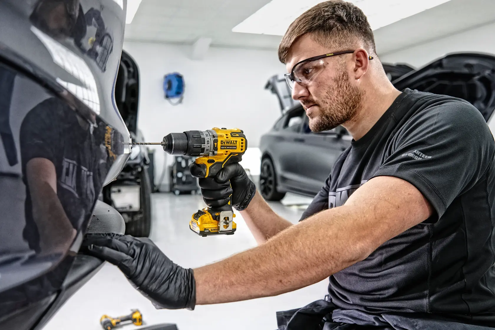 Eine Person mit schwarzen Handschuhen verwendet einen DEWALT Akkubohrer am hinteren Stoßfänger eines Autos in einer Werkstatt. Das Gesicht der Person ist aus Datenschutzgründen unkenntlich gemacht.