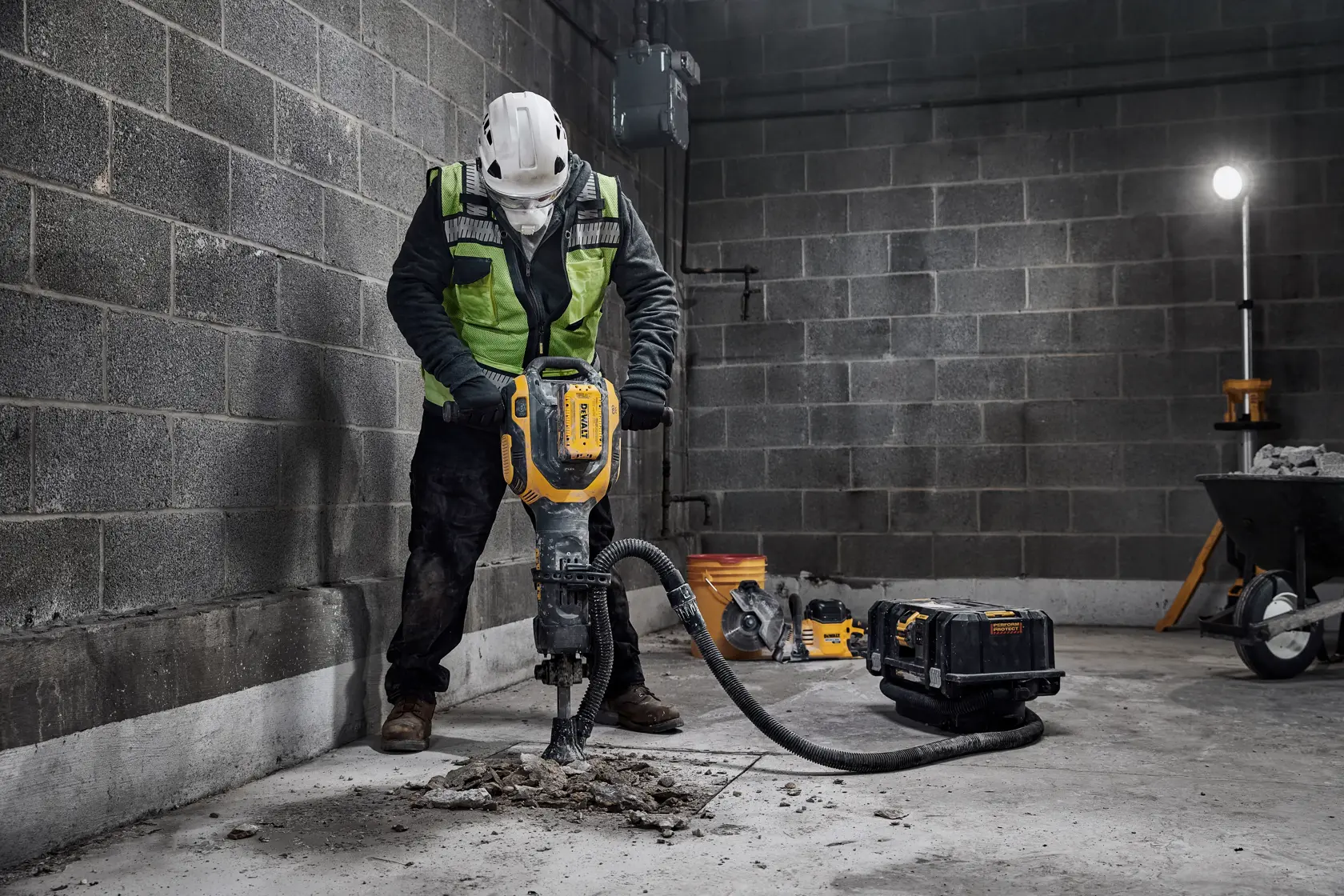 A construction worker in safety gear operates a DEWALT electric jackhammer to break up concrete on a worksite. The tool is connected to a vacuum system and there is construction equipment in the background.
