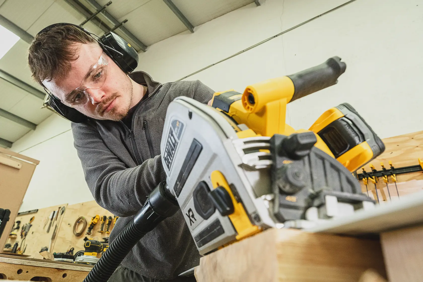 Person using a DEWALT cordless circular saw on wood, wearing safety glasses and ear protection in a workshop.