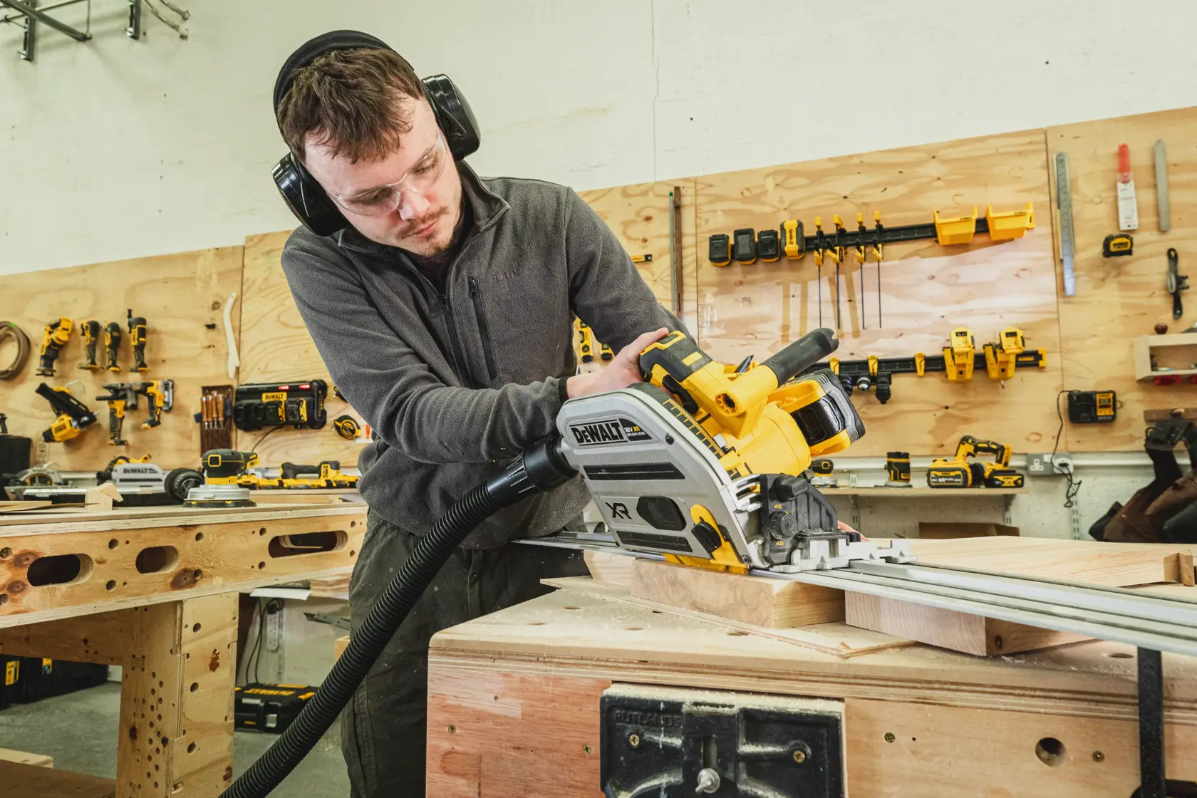 Person using a DEWALT cordless track saw on wood in a workshop, wearing ear and eye protection.