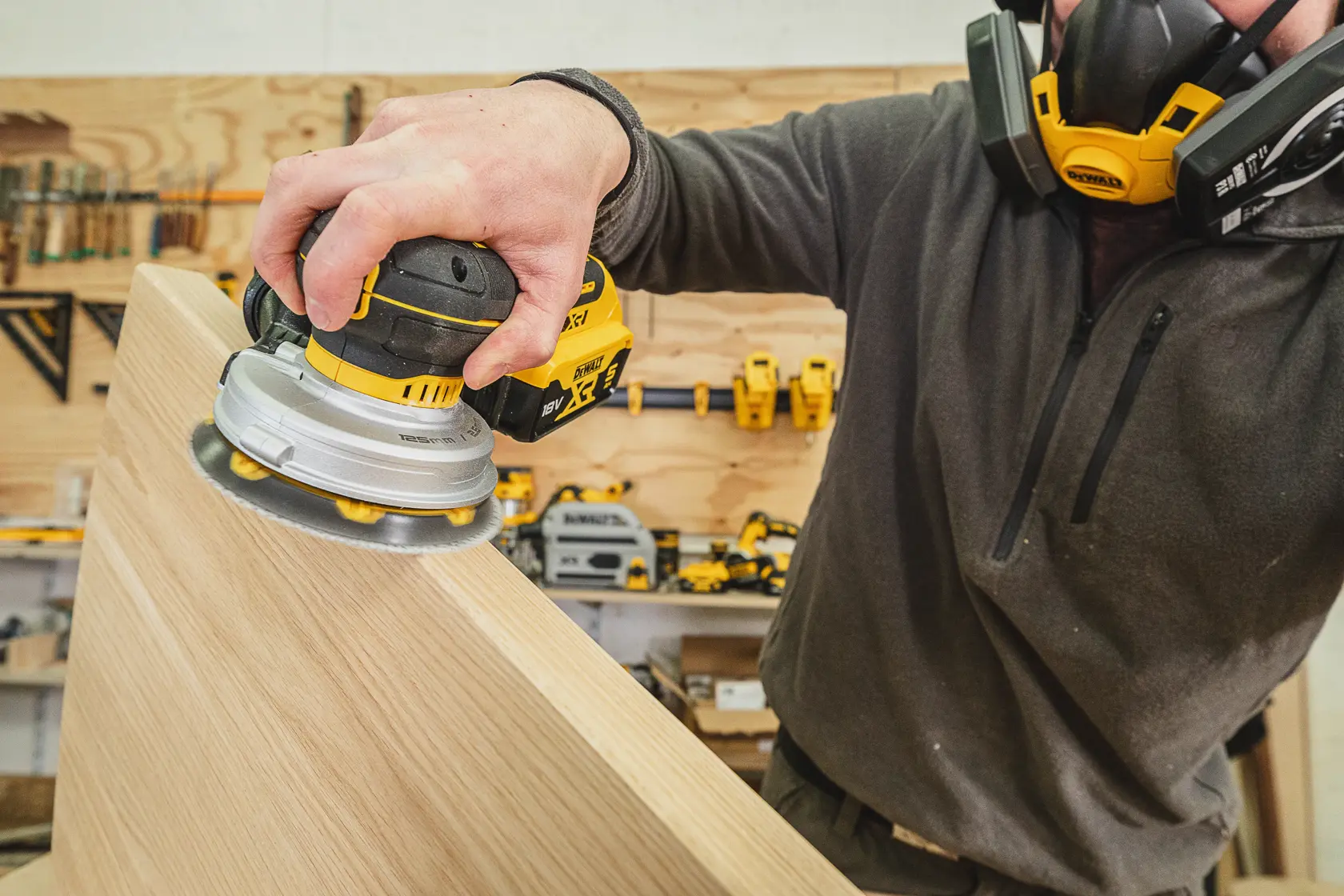 Person uses a DEWALT cordless sander on a wooden surface in a workshop, wearing a protective mask.