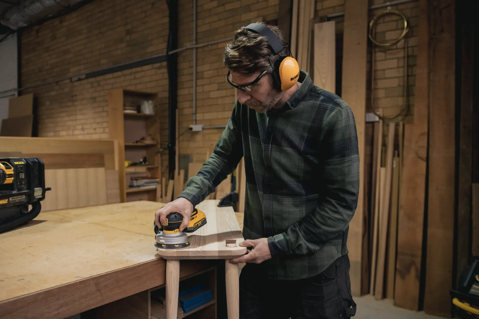 Person using a DEWALT cordless sander on wood in a workshop while wearing ear and eye protection.