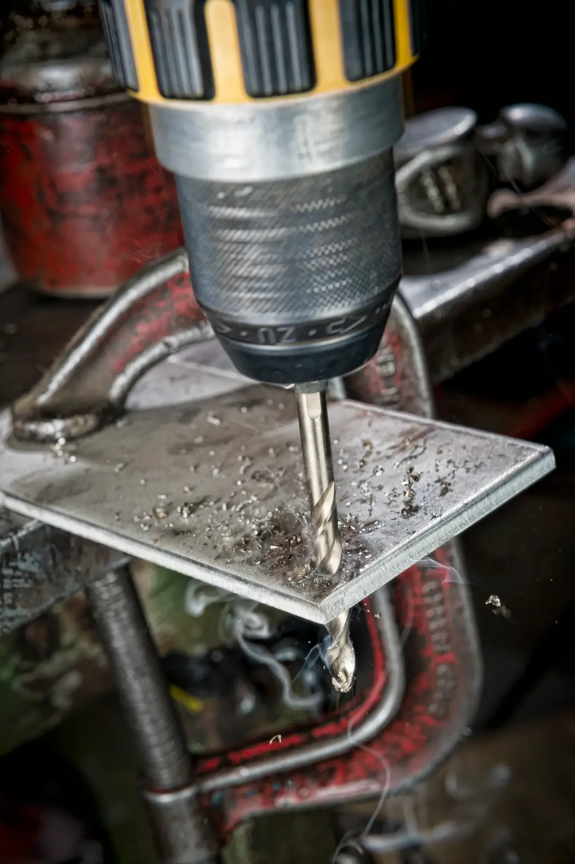 Close-up of a drill creating a hole in a metal plate, held in place by a clamp, with metal shavings visible.