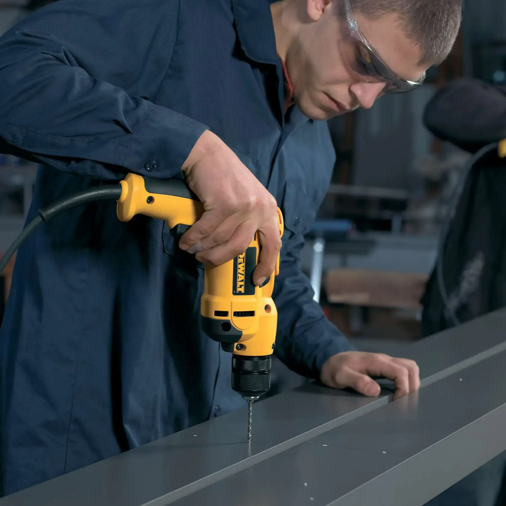 Person drilling metal with a yellow DEWALT electric drill in a workshop.