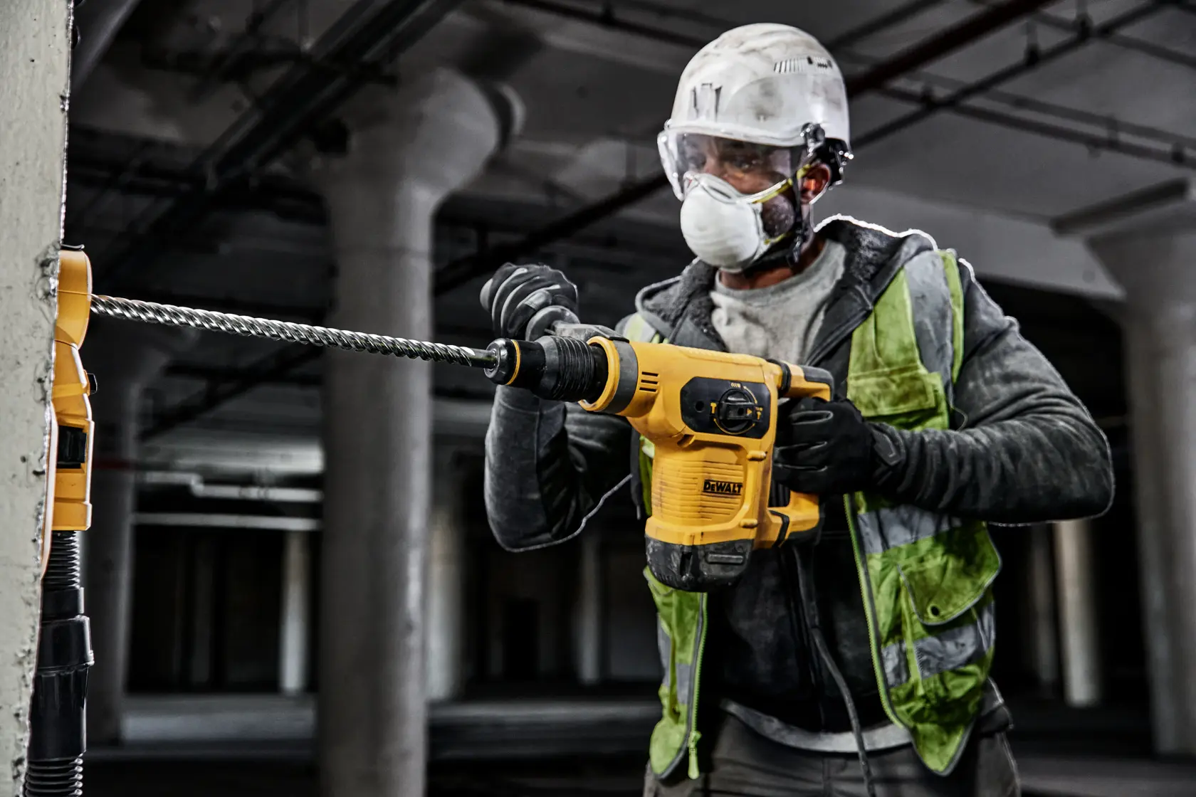Trabajador con chaleco y casco usa taladro martillo DEWALT amarillo en pared de concreto en sitio industrial.