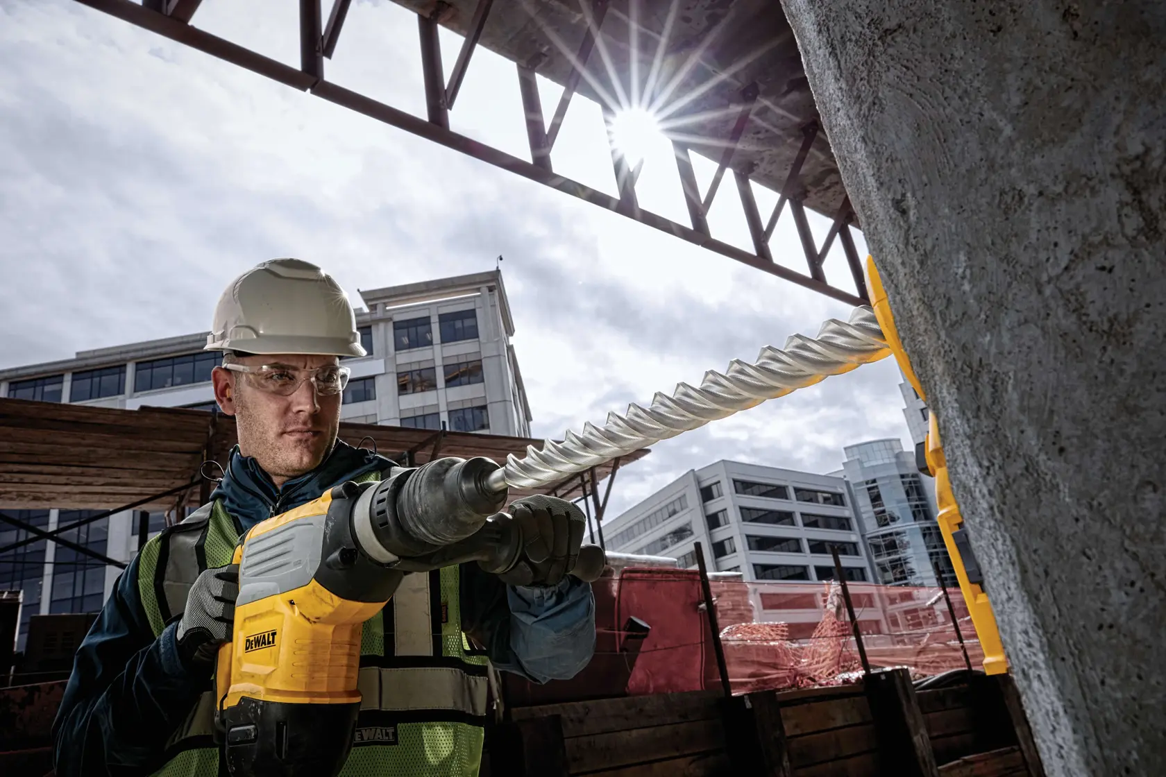 Un obrero de la construcción utiliza un martillo perforador DEWALT con una broca larga para taladrar concreto en una obra al aire libre. El trabajador lleva equipo de seguridad, incluyendo casco y chaleco reflectante. Al fondo se ven edificios modernos.