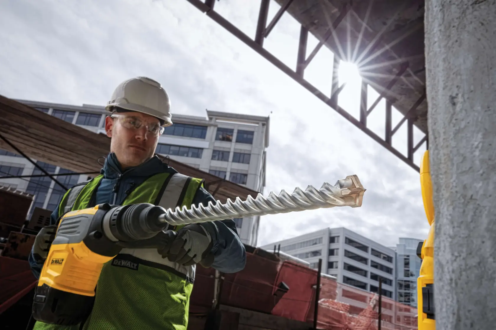 Trabajador con martillo rotativo DEWALT y broca DT60847 en obra, edificios al fondo.