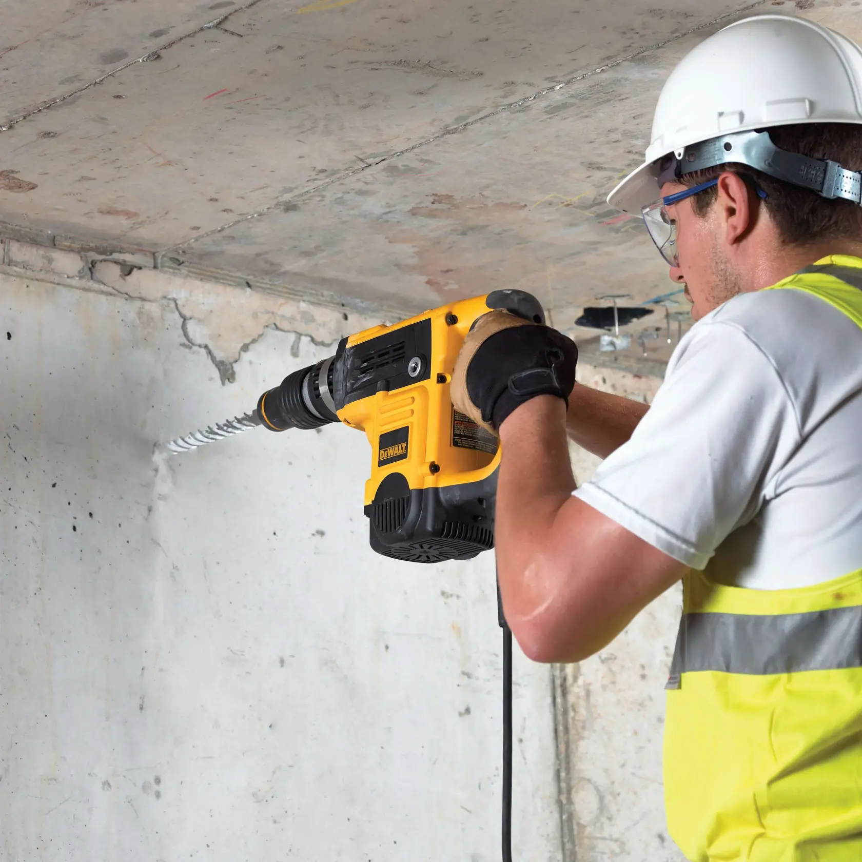Arbeiter bohrt Betonwand mit gelbem DEWALT Werkzeug, trägt Schutzkleidung und Handschuhe.