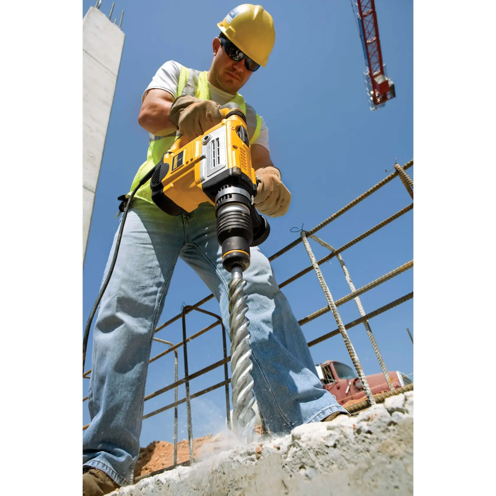 Worker using a DEWALT hammer drill on concrete at a construction site, wearing safety gear and gloves.
