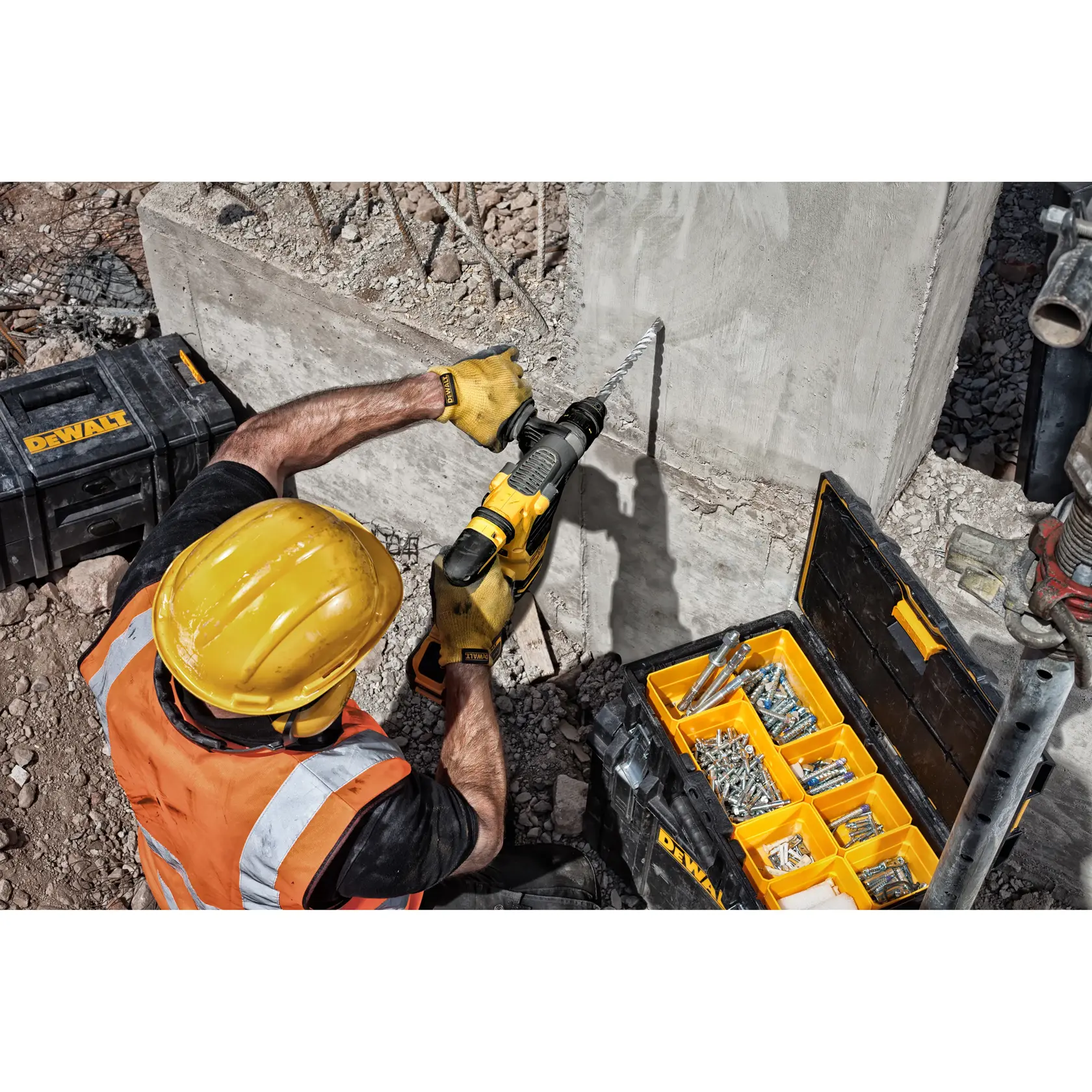 Trabajador usa taladro DEWALT en pared de hormigón junto a caja de herramientas abierta con tornillos en obra.