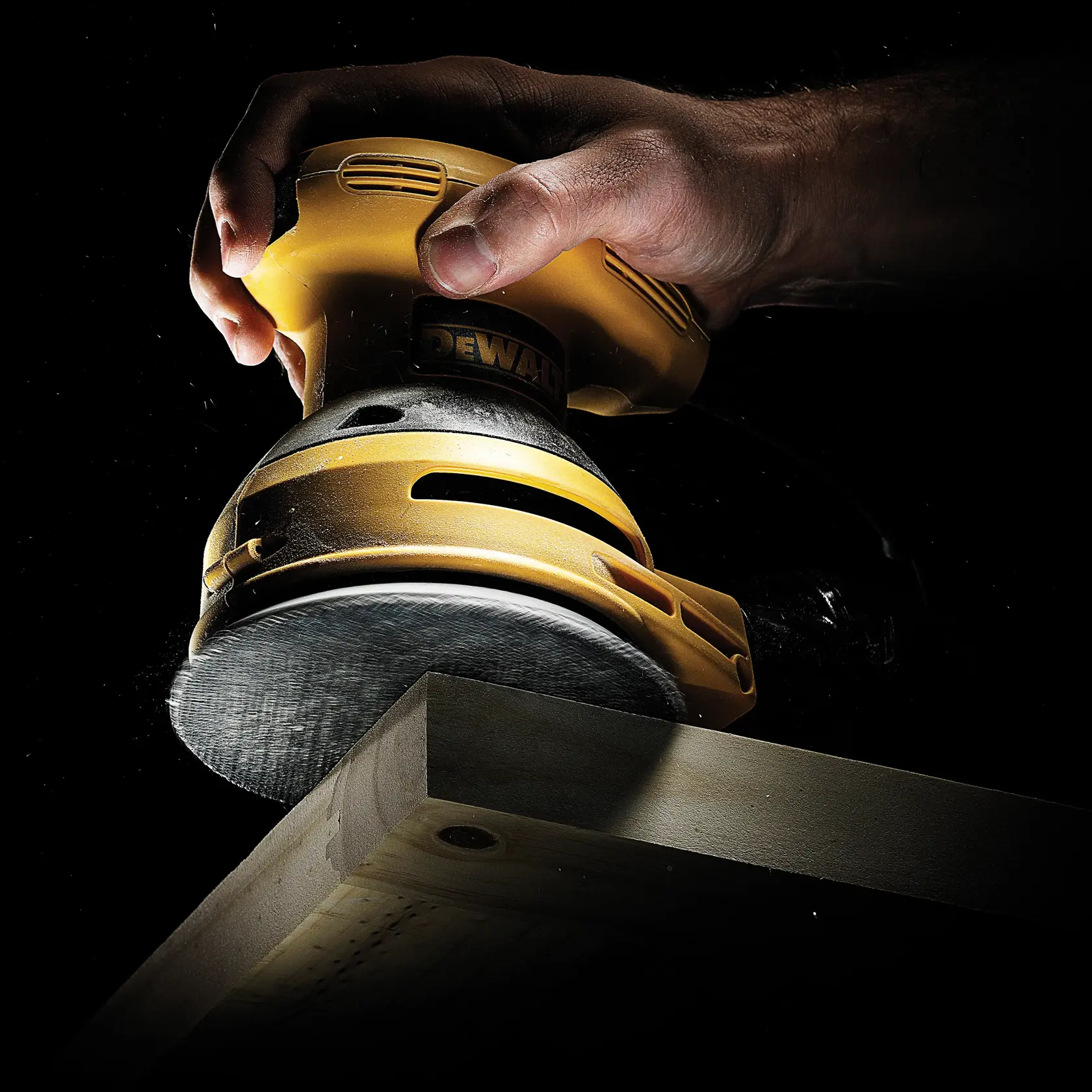 Hand using a DEWALT sander to smooth a wooden plank edge in a dark setting.
