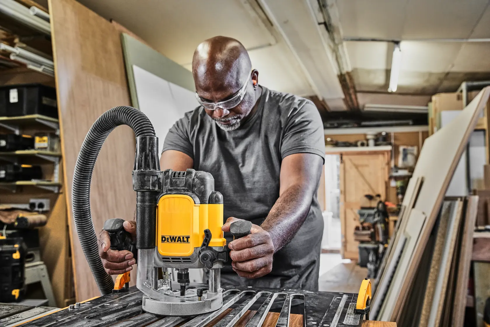 A person using a DEWALT DWE625 router tool in a woodworking workshop. The tool is placed on a workbench with wood pieces and a vacuum hose attached. The workshop is filled with various materials and equipment.